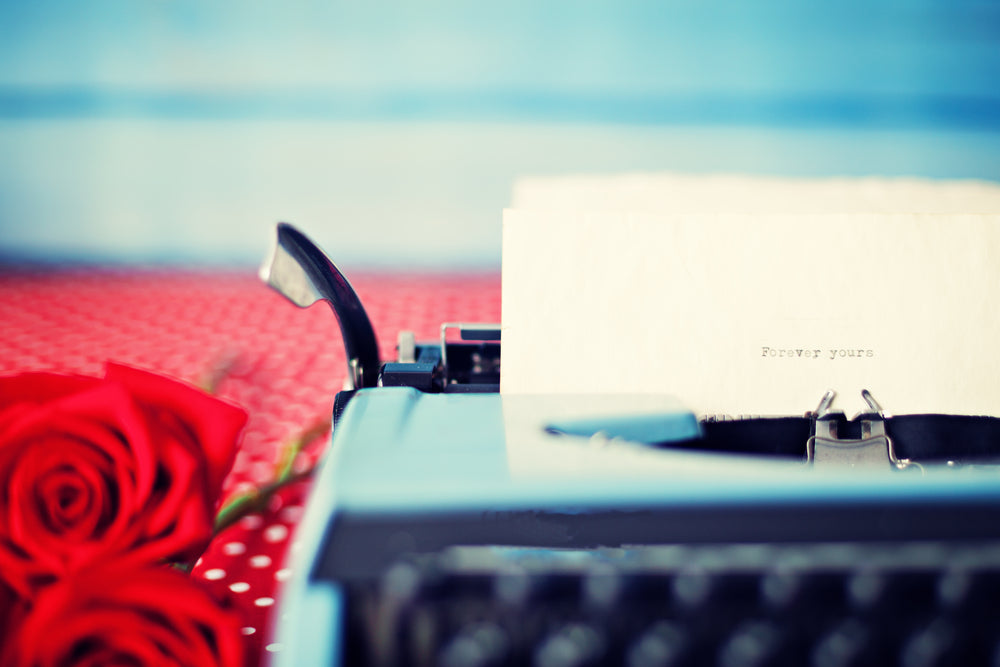 Typewriter with "Forever yours" typed on a page, a red rose resting on a polka-dot cloth beside it.
